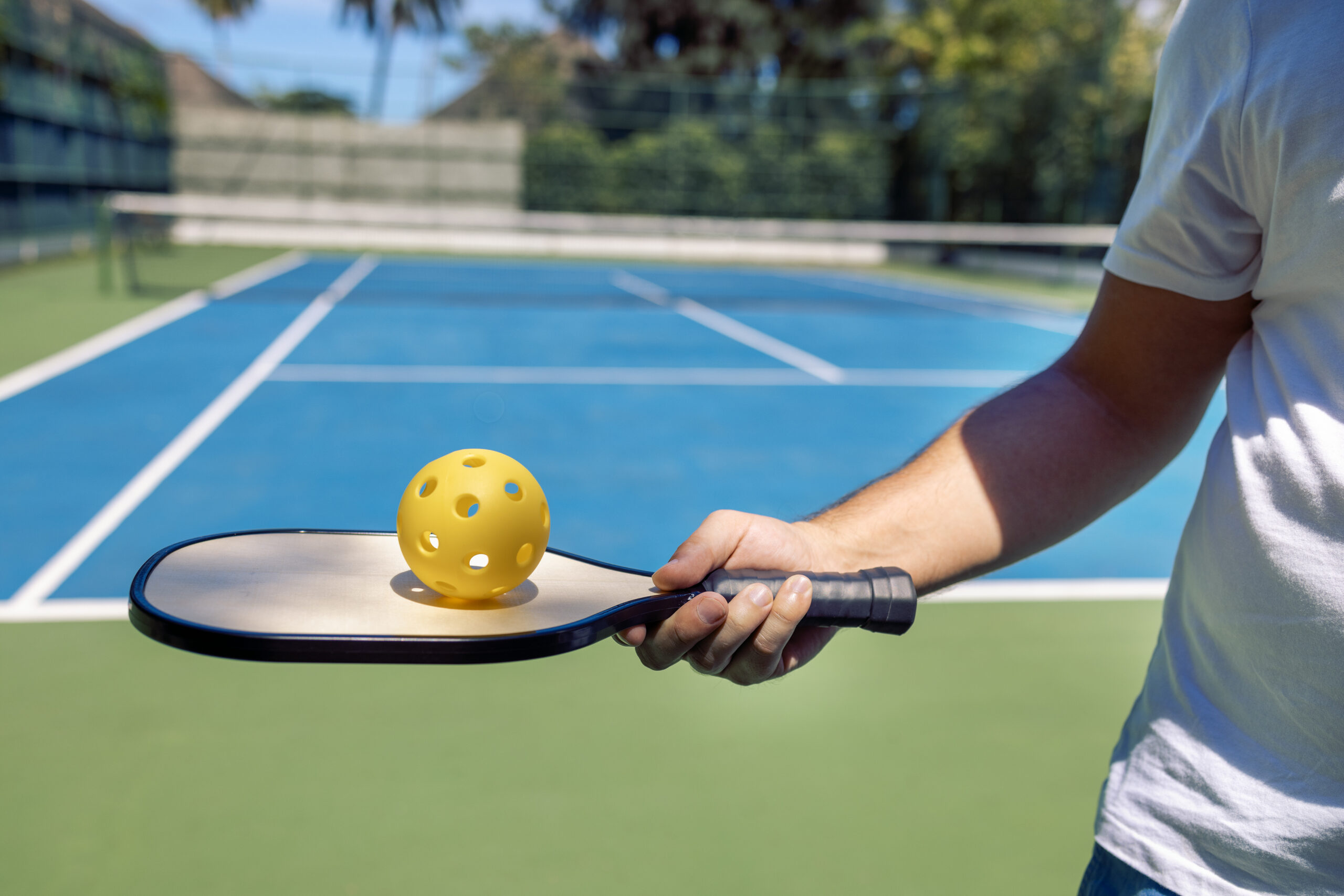 This image shows someone playing pickleball on what appears to be a dedicated pickleball court. The person is holding a pickleball paddle with a yellow pickleball (recognizable by its distinctive holes) balanced on the paddle face. The court surface is blue with white lines marking the boundaries and zones typical of pickleball courts. In the background, you can see green space and trees, suggesting this is an outdoor recreational facility. The lighting and setting give it a pleasant, sunny day atmosphere perfect for outdoor sports.