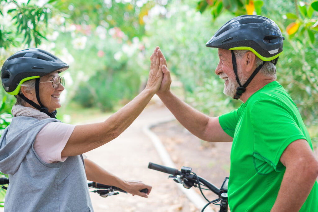 Active senior couple giving high-five while biking outdoors, wearing matching gray helmets with green accents on tree-lined path