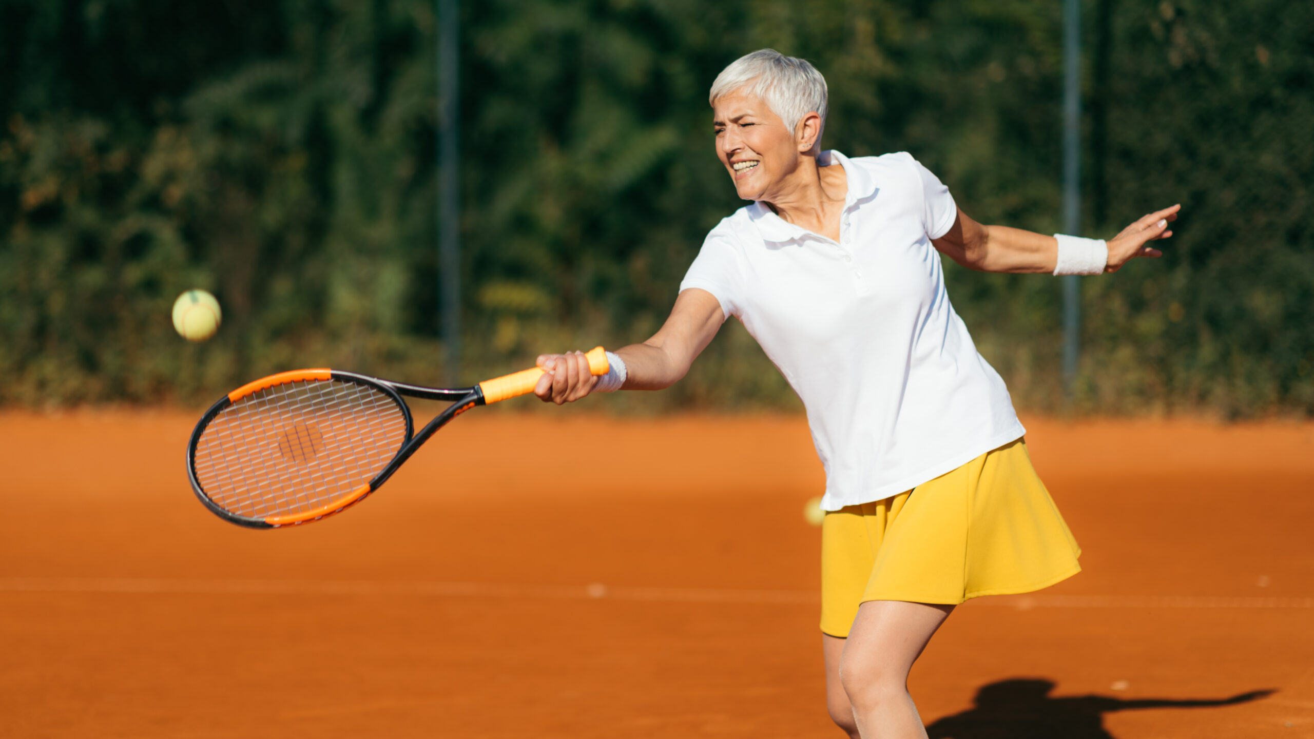 This image shows a mature woman with short gray hair playing tennis on a clay court. She's wearing a white polo shirt and bright yellow tennis skirt, and is captured mid-swing with a tennis racket as she reaches to hit a tennis ball that's visible in the frame. Her expression shows focus and enjoyment as she plays. The distinctive reddish-orange clay court surface is clearly visible, with green trees and foliage creating a natural backdrop. The lighting suggests it's during golden hour, creating warm, pleasant conditions for outdoor tennis.RetryClaude can make mistakes. Please double-check responses.